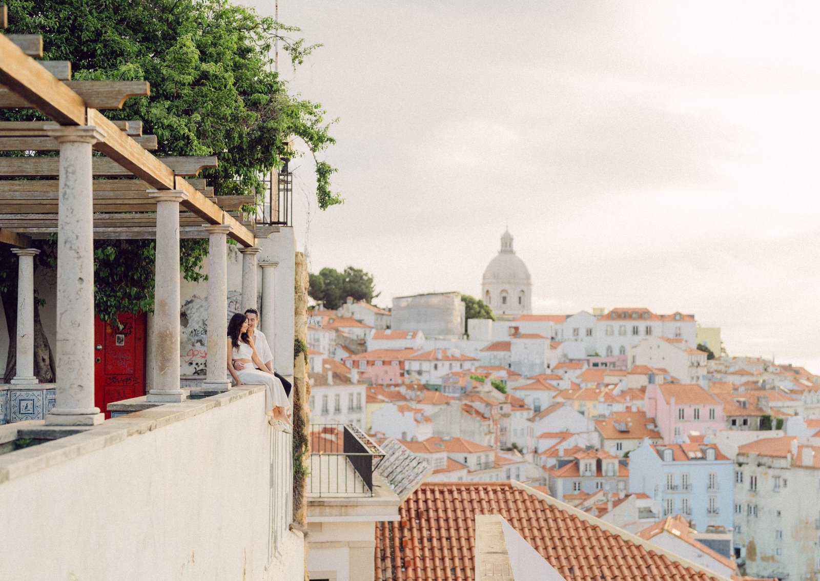 Couple photographed at a Lisbon viewpoint overlooking the Alfama rooftops at golden hour — engagement photographer Flavia Grandi, Lisbon Portugal