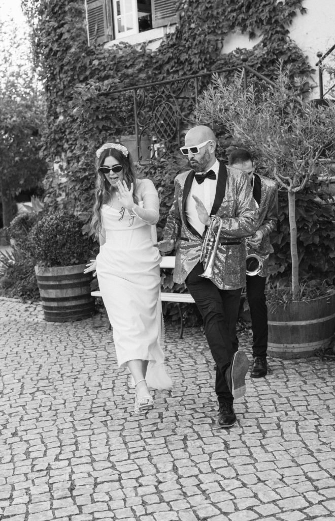 Black and white photo of a bride in sunglasses walking on cobblestones alongside a saxophonist in a gold sequin jacket