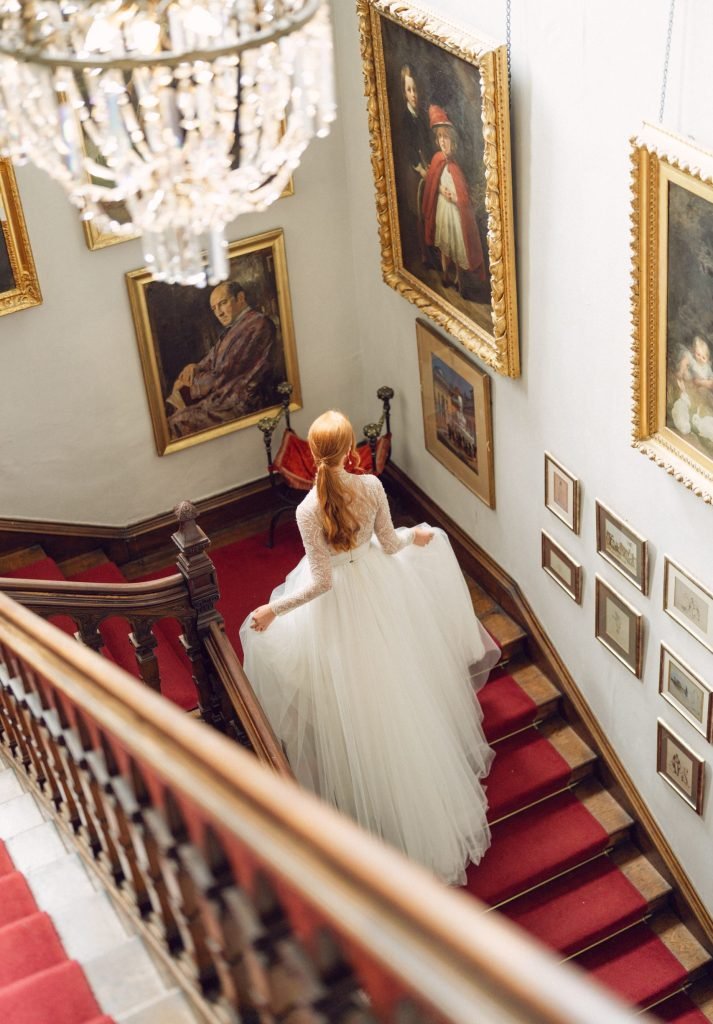 Overhead view of a bride in a full tulle gown descending a grand red-carpeted staircase lined with oil paintings and a crystal chandelier