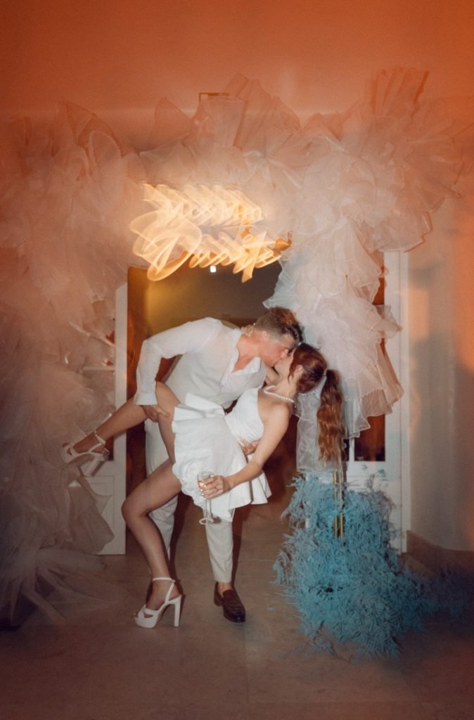Bride and groom kissing dramatically in a warmly lit doorway, surrounded by billowing white fabric and teal feathers, with a glowing neon sign above