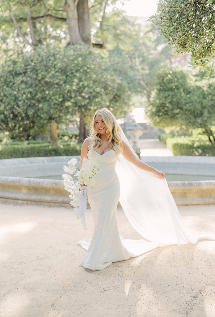 Bride in a flowing white gown holding a white bouquet and lifting her veil, standing near an ornamental fountain in a lush garden