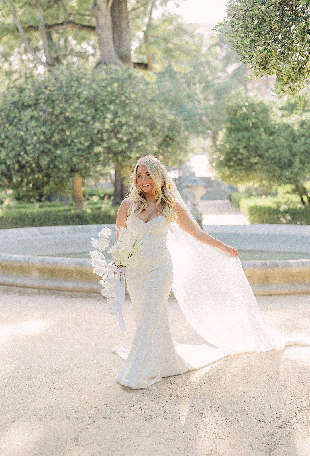 Bride in a flowing white gown holding a white bouquet and lifting her veil, standing near an ornamental fountain in a lush garden